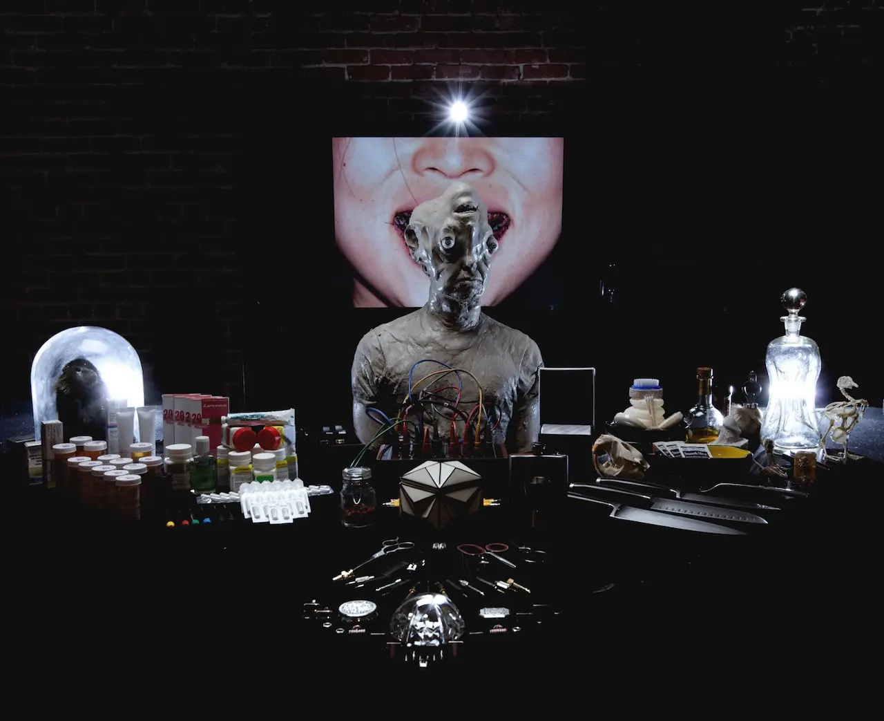 Staged still life: a clay-headed figure at a table of bottles, pills, knives and curios, an open mouth on a screen behind