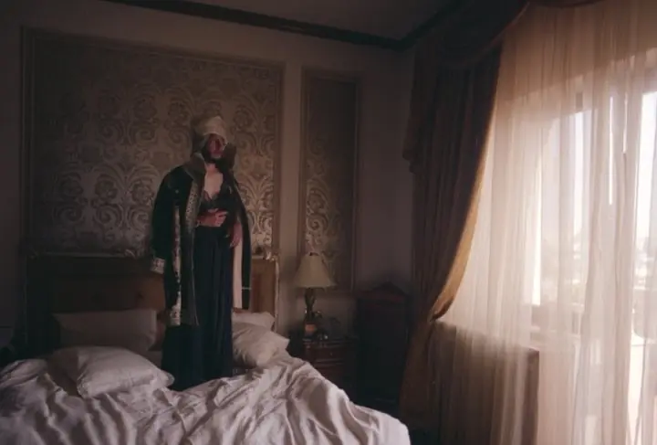 A figure in a long robe standing on an unmade bed in an ornate room