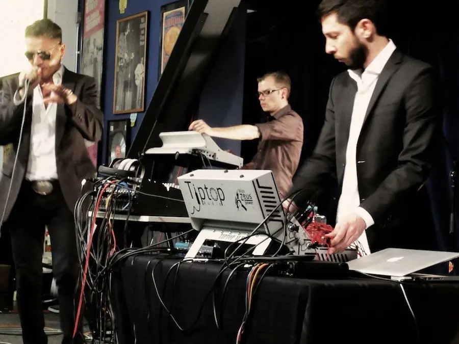 Three performers on stage: a singer in sunglasses, and two at a modular case and a laptop