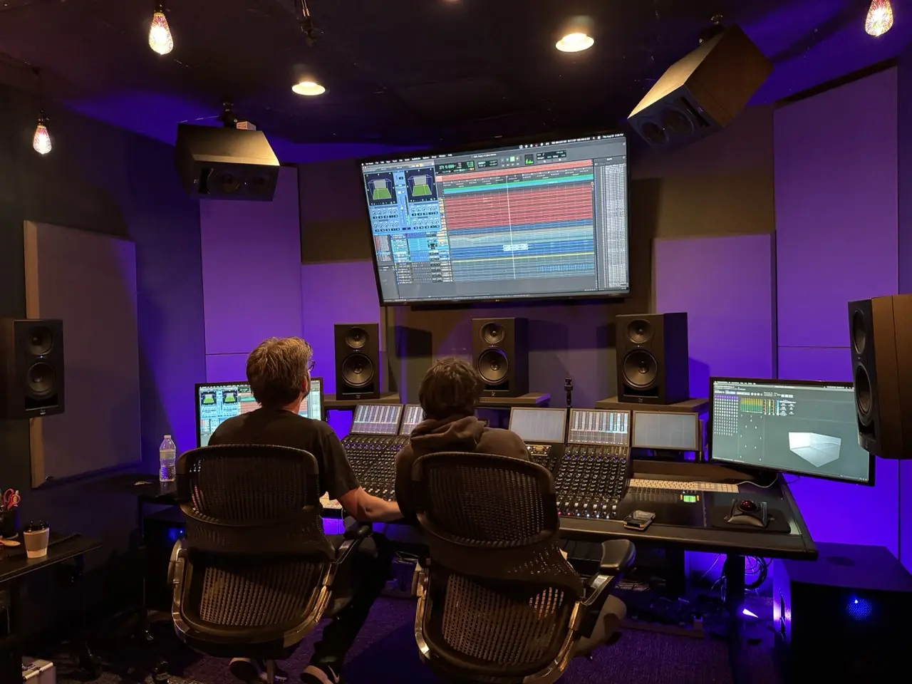 Two people at a mixing desk under large screens in a purple-lit studio