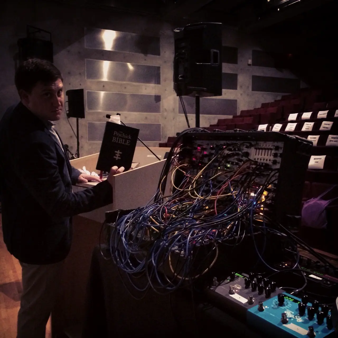 MOCA, before the show: a man at a lectern holding The Psychick Bible beside a patched modular rig, the theatre seats behind
