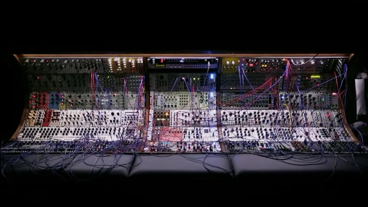 A wall of Eurorack modular synthesizer cases, fully patched, lit against a dark room