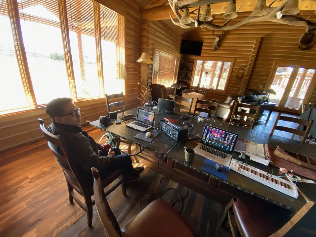 A log-cabin studio with tall windows, a person at the desk among synths