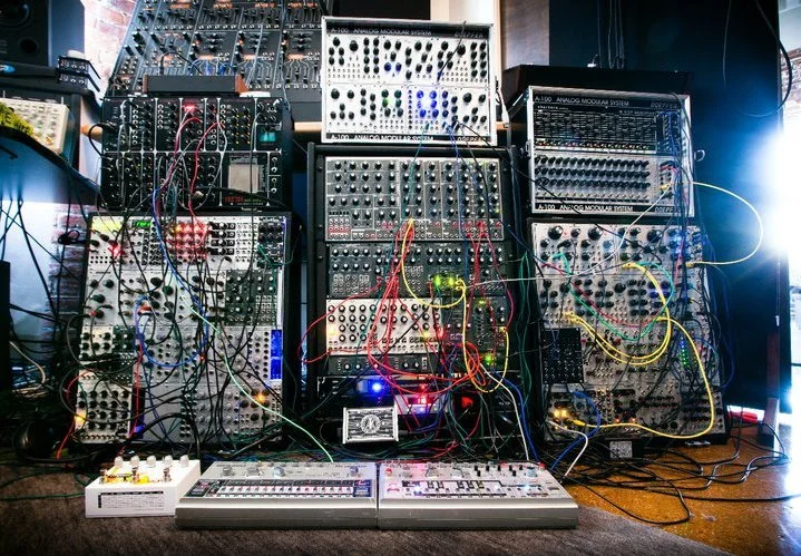 The Cyrusrex studio rig: stacked cases of Eurorack modules with a Devil Fish TB-303 and a TR-606 on the floor in front, lit in blue, with patch cables everywhere