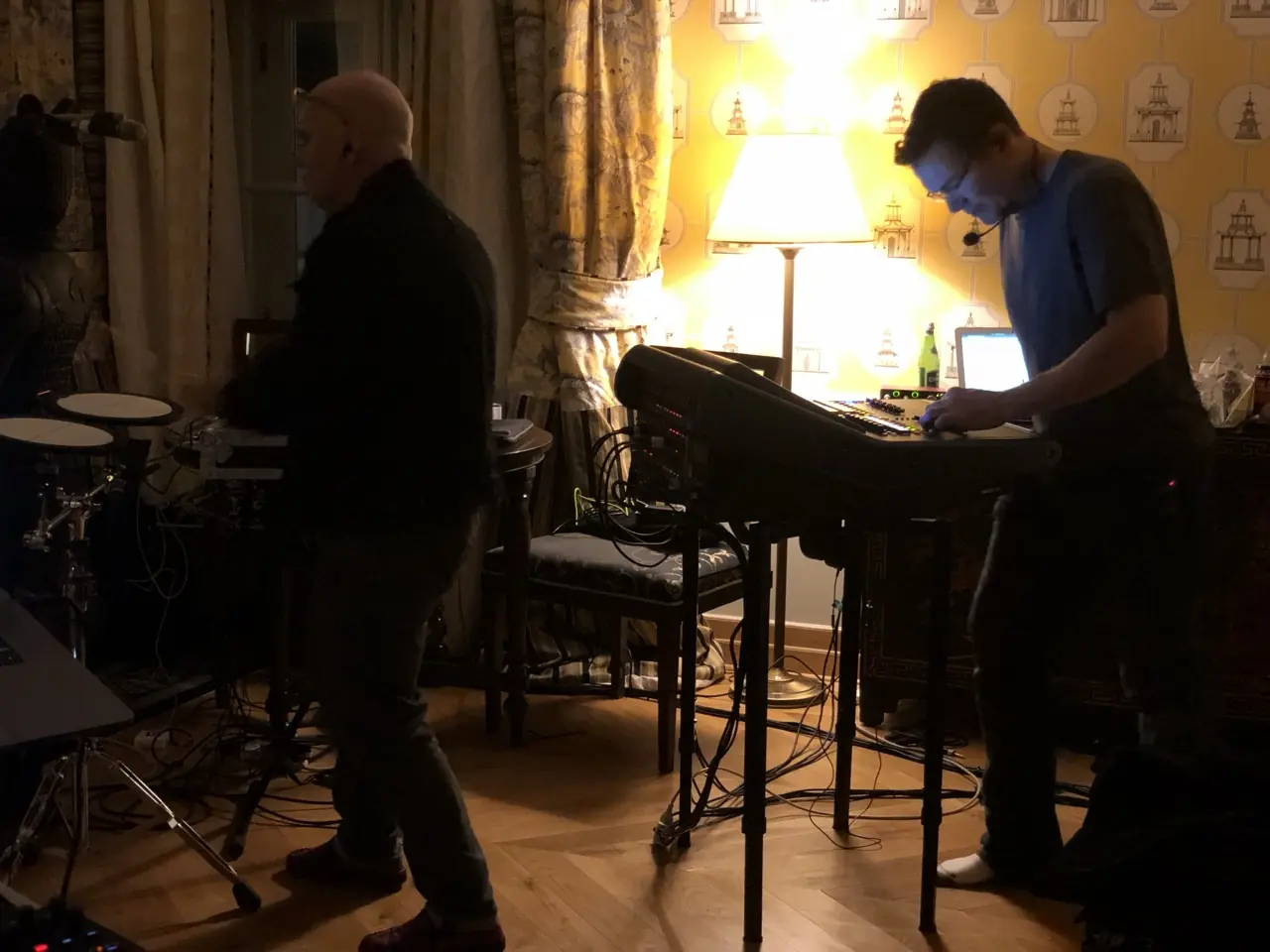 Two men with keyboards and drum pads in a room with patterned wallpaper