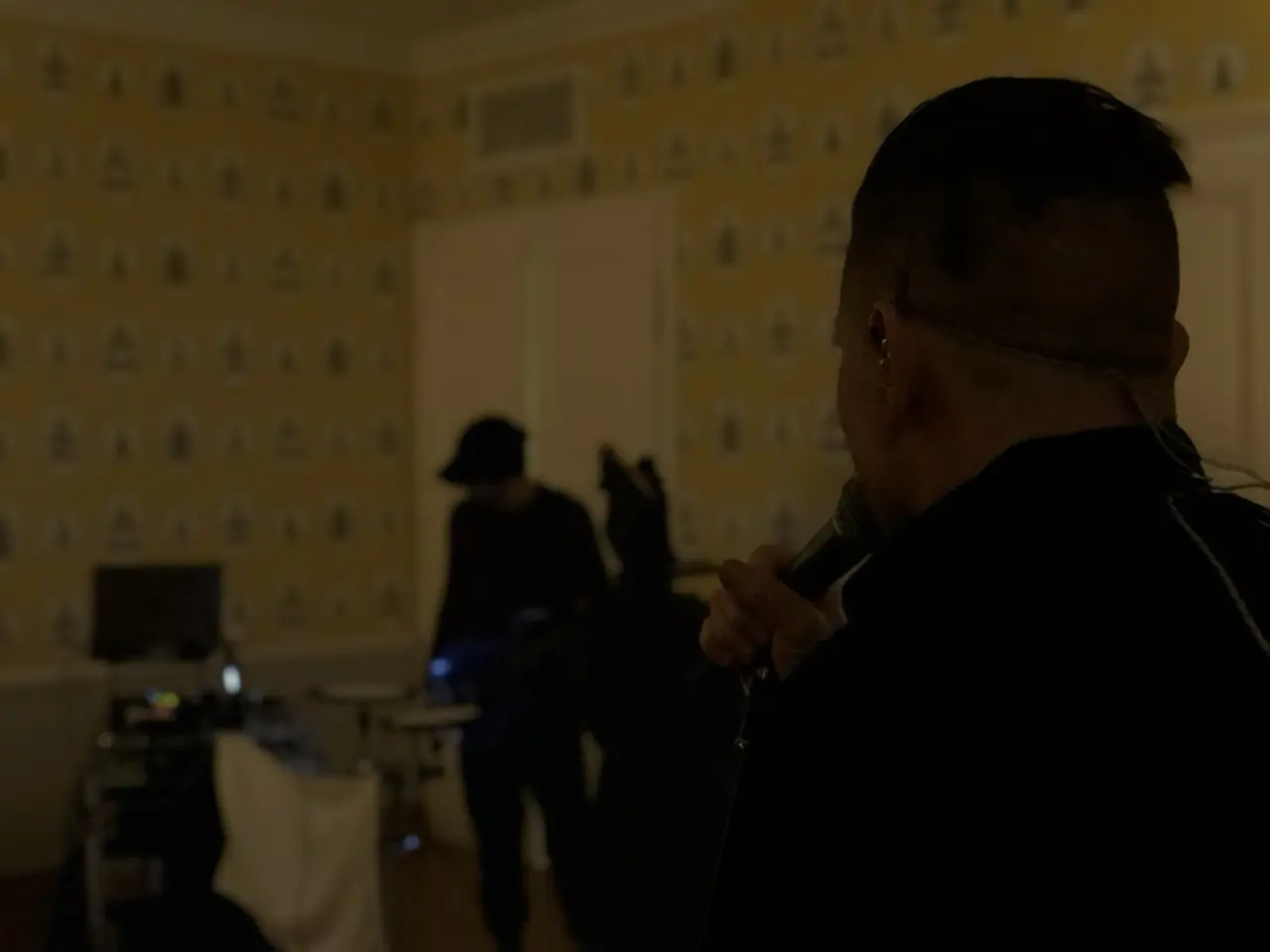 A man with a microphone in a dim room, others setting up behind