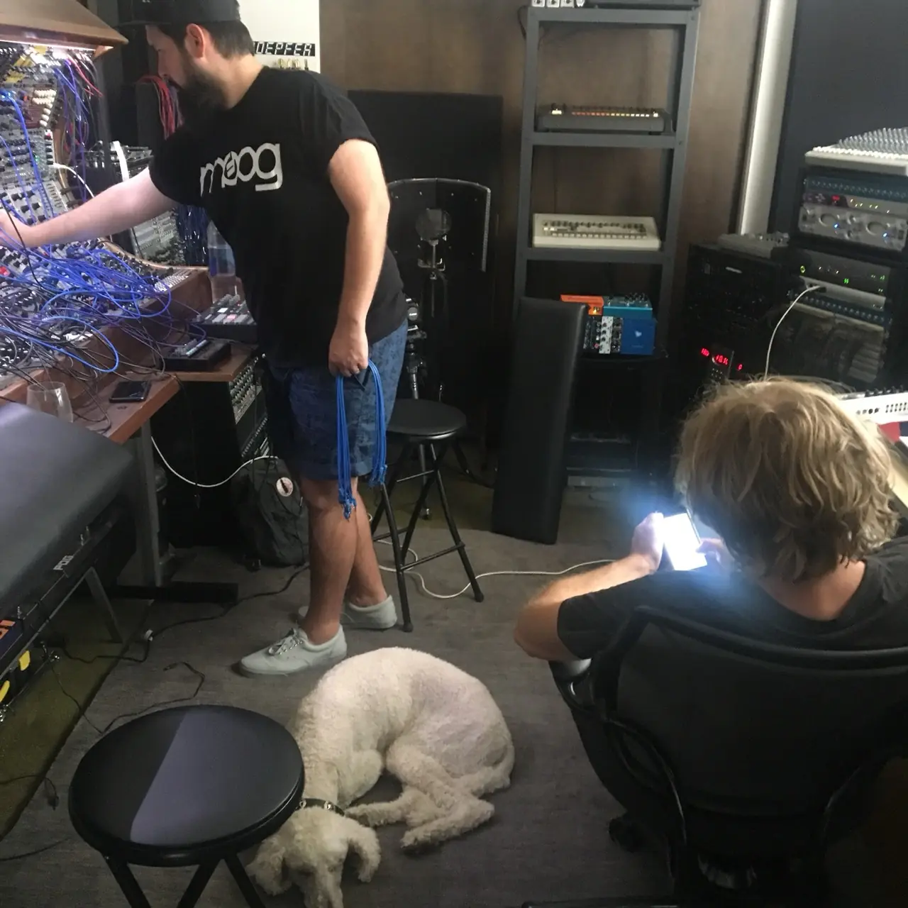 A man at a modular synth and another on the floor with a phone, a white dog asleep beside them