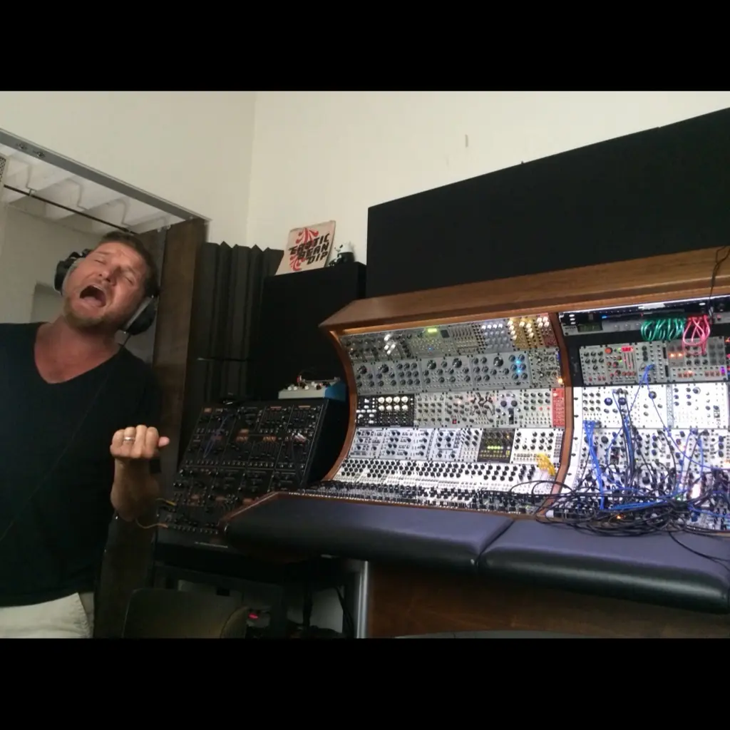 A man in headphones singing hard in front of a modular synth